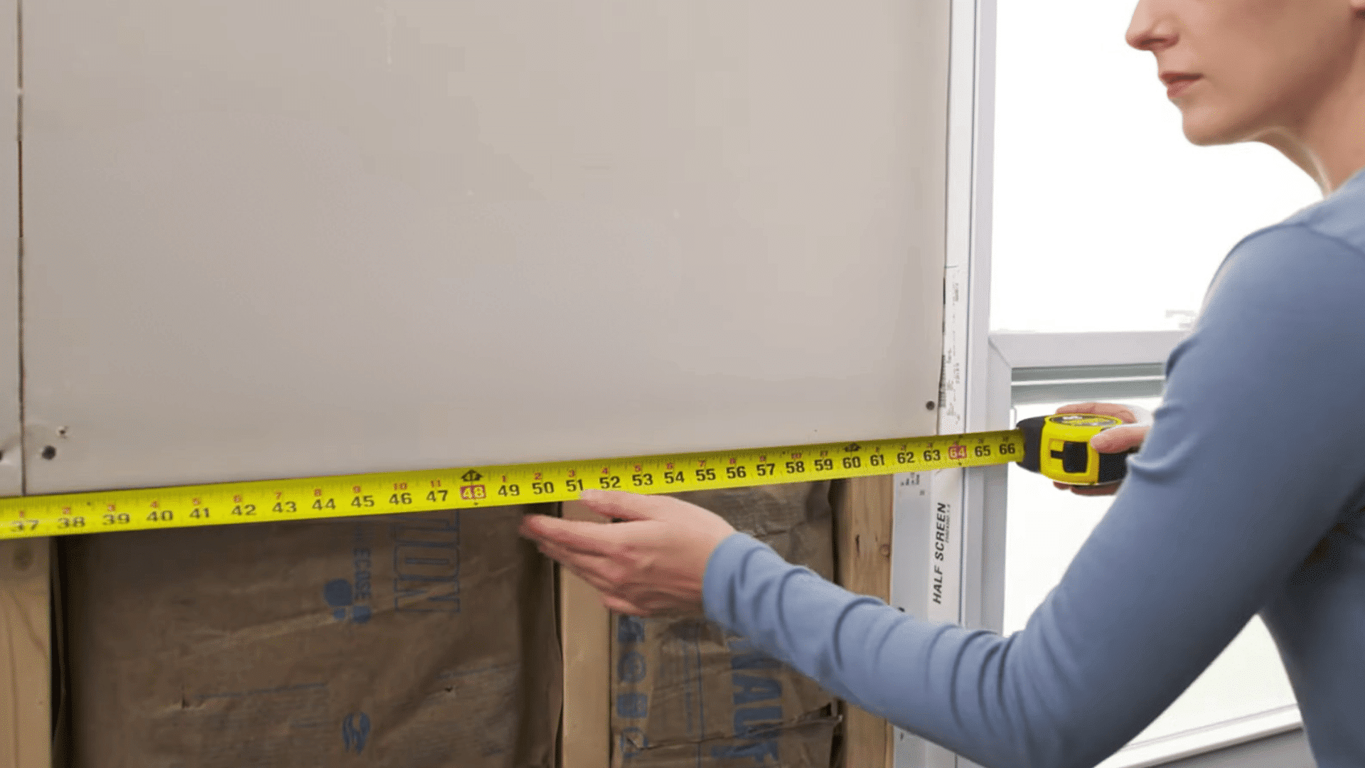 woman measuring drywall panel width with a tape measure during the first step of a drywall installation project