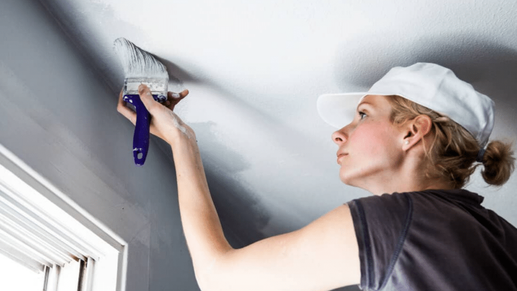 How to Easily Paint a Popcorn Ceiling as a Beginner? woman painting along the ceiling edge with a brush while creating a clean line between the wall and ceiling surface