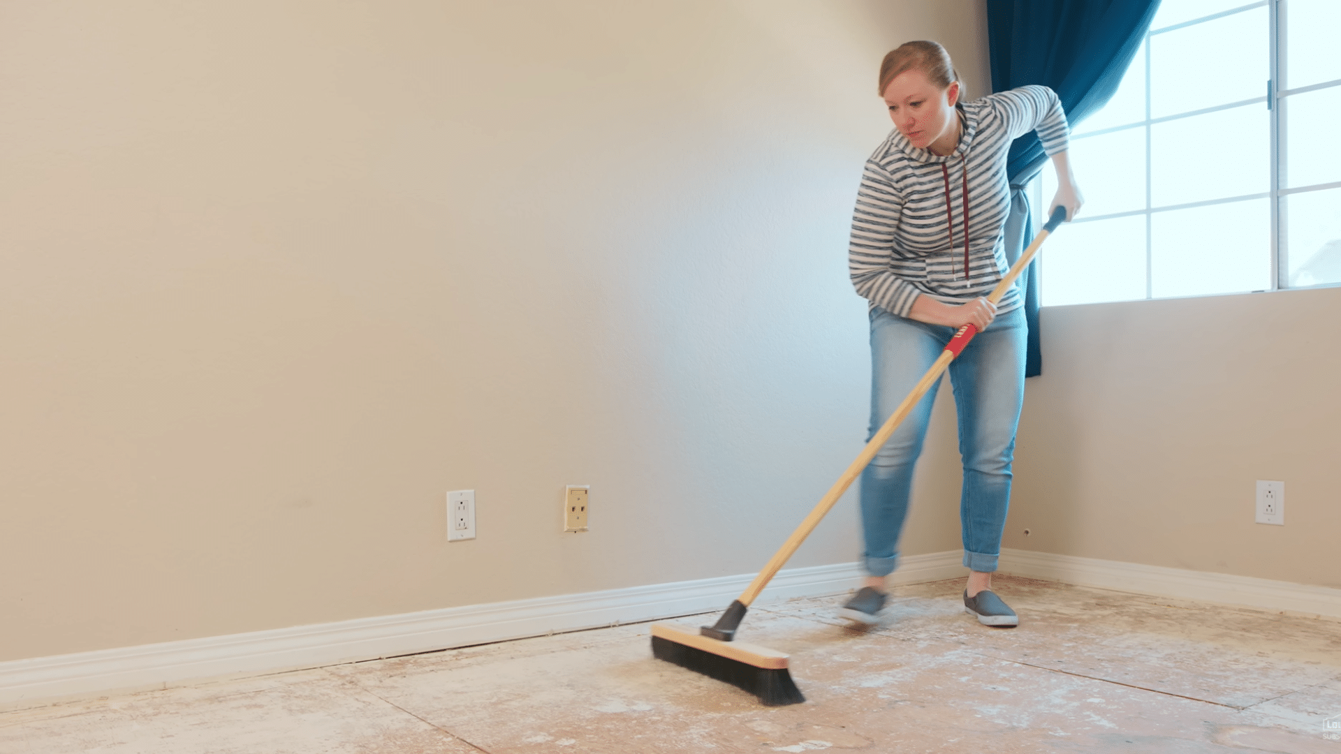 woman sweeps dust and debris from an exposed plywood subfloor in an empty room before flooring installation begins