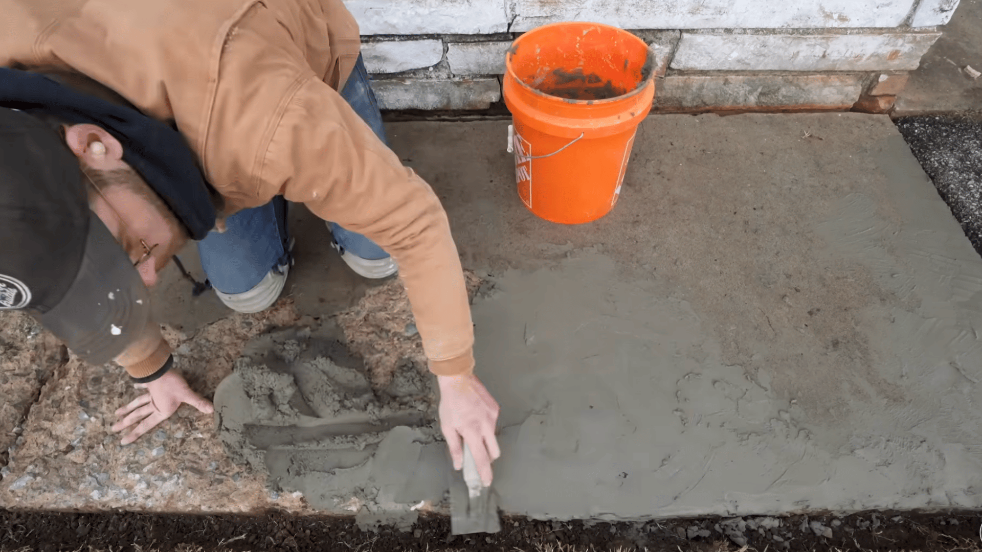 worker applying fresh concrete resurfacer to repair a spalled sidewalk section beside a building foundation wall