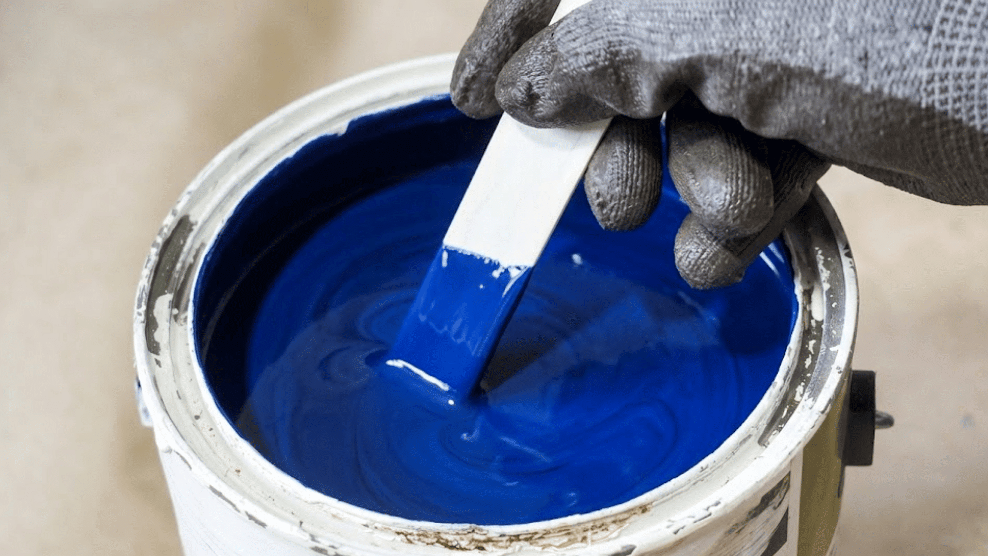 a gloved hand stirs bright blue paint with a mixing stick inside an open paint bucket preparing paint for application