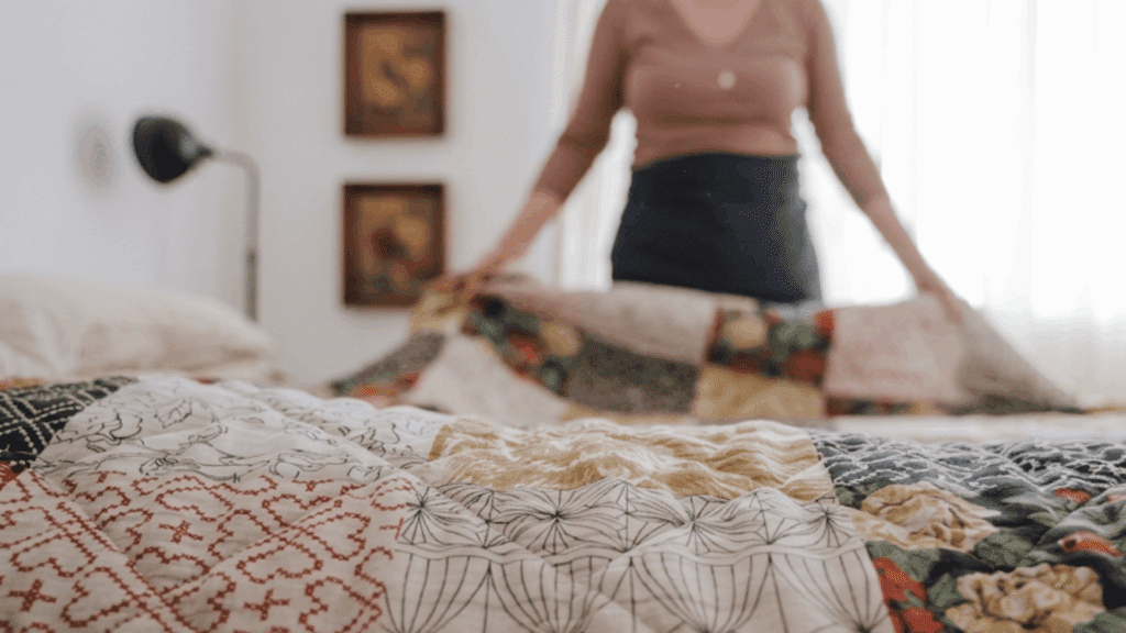 a person spreads a colorful patchwork quilt across a neatly arranged bed in a bright and cozy bedroom