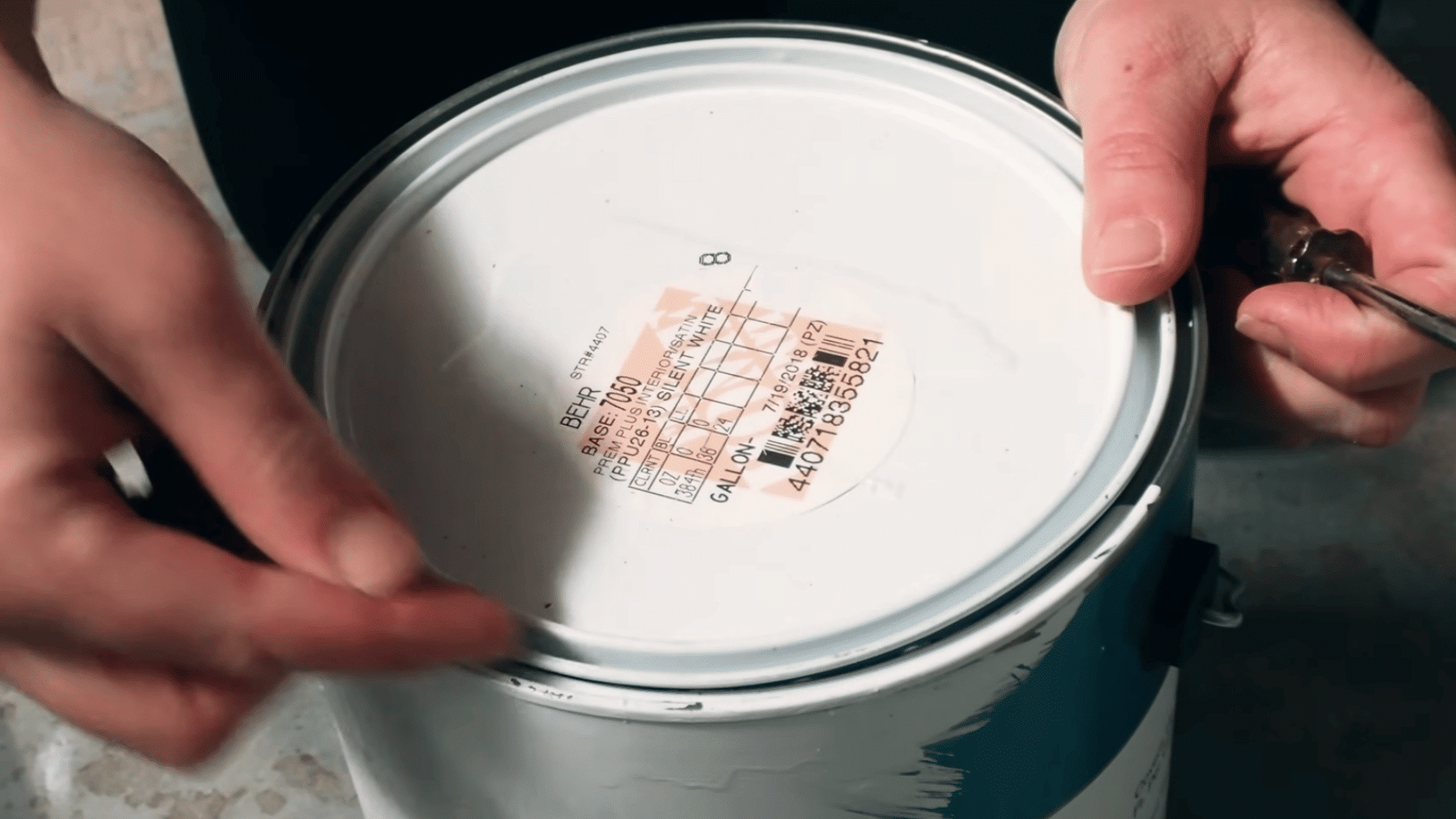 close up of hands inserting a flathead screwdriver beneath the rim of a sealed paint can lid