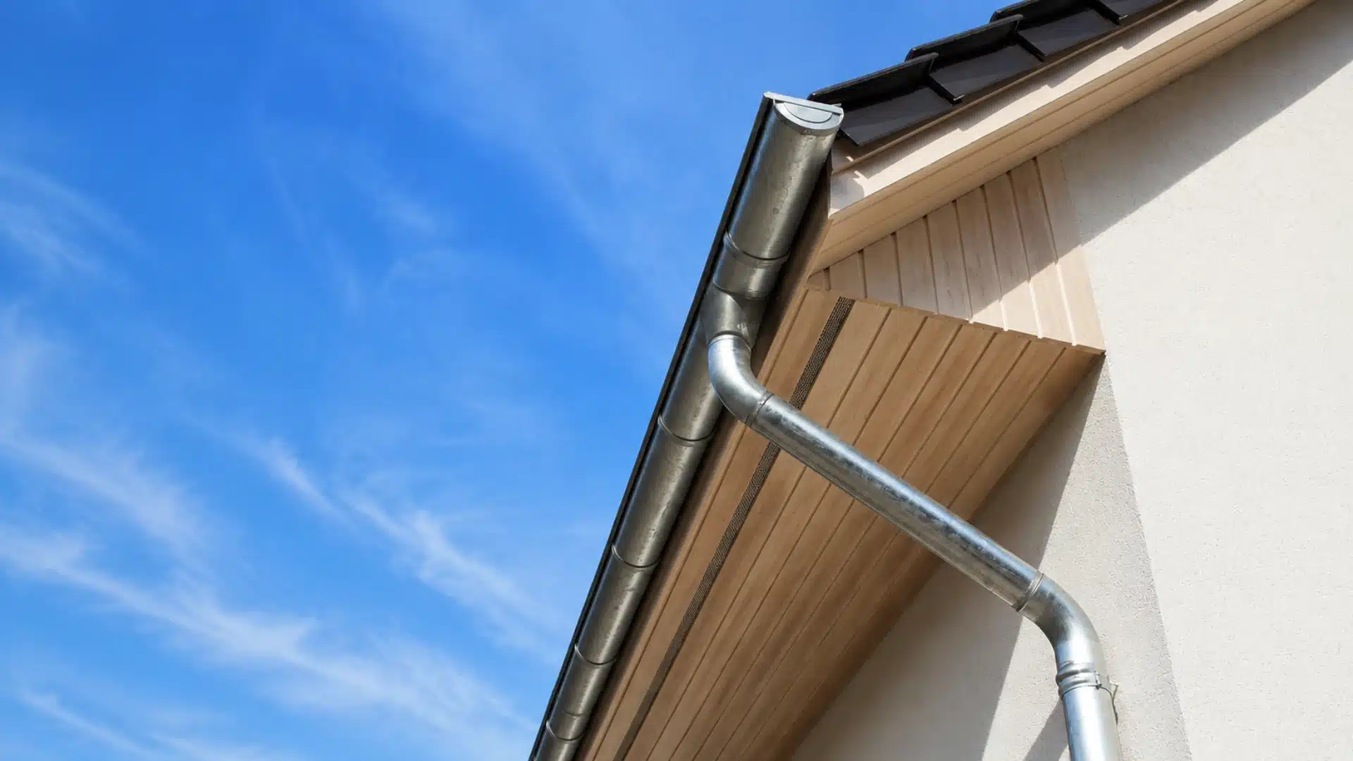 Modern house eave with wooden soffit, metal gutter, and downspout against a blue sky.