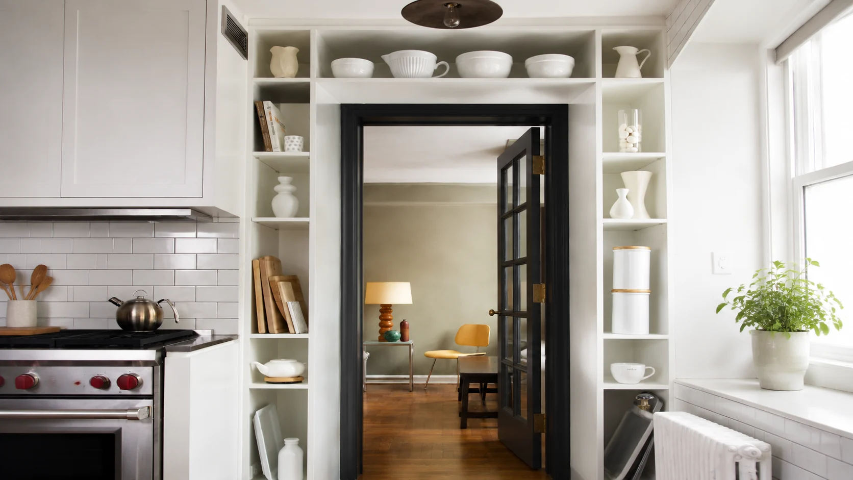 White kitchen doorway with built-in open shelves displaying ceramic dishes, vases, books, and minimalist decor around a dark framed door.