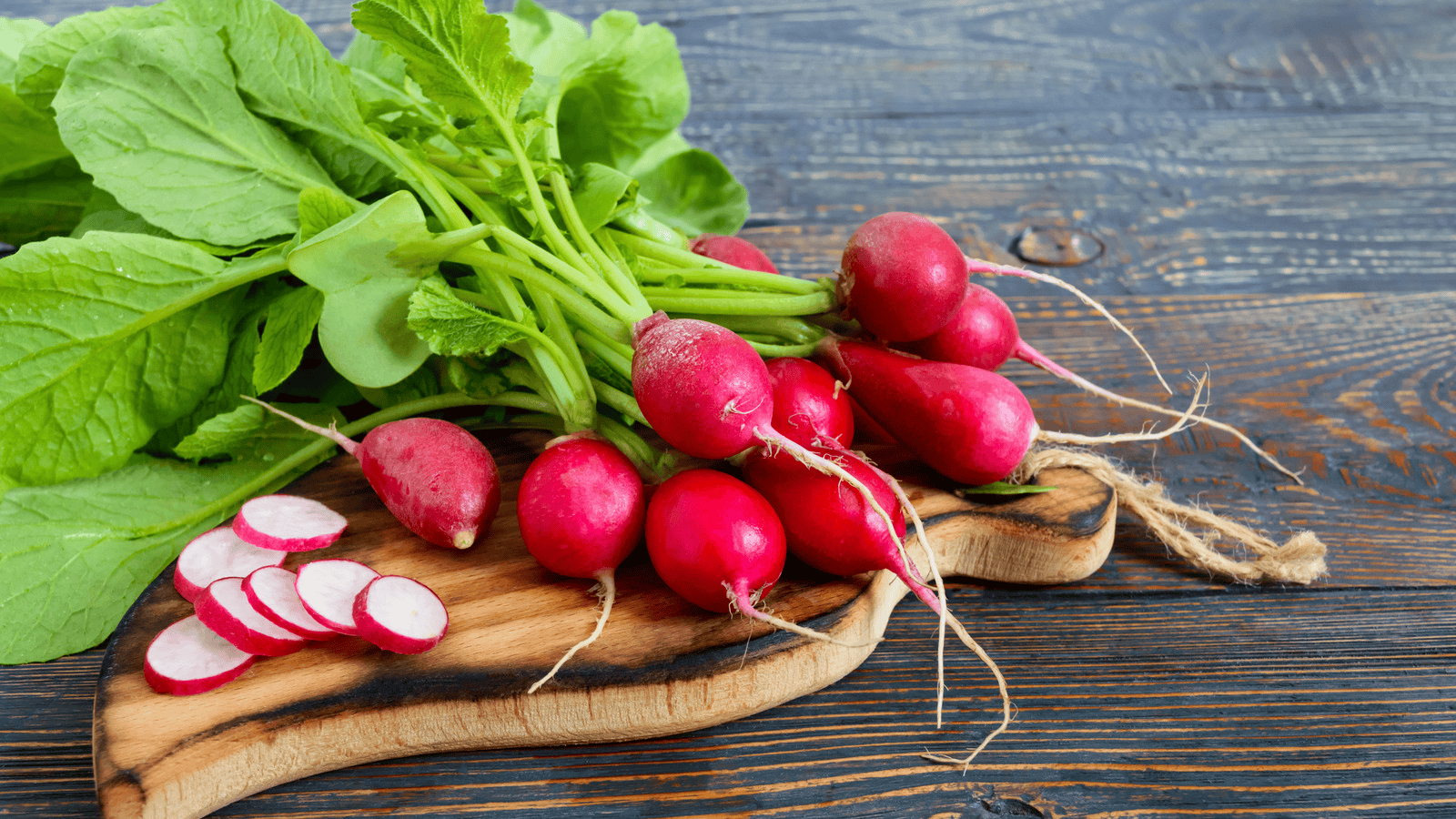 fresh red radishes with leafy tops and sliced pieces arranged on a wooden board over a rustic wooden table