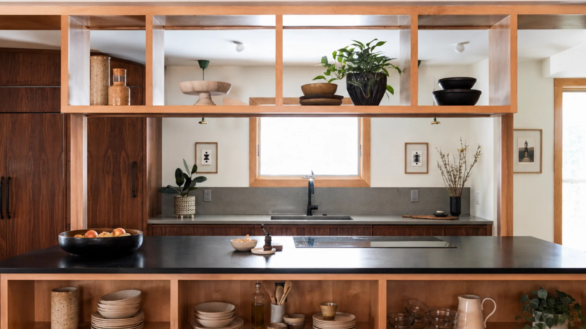 Warm wood kitchen with open island shelving, stacked dishes, vinyl records, black counters, and indoor plants.