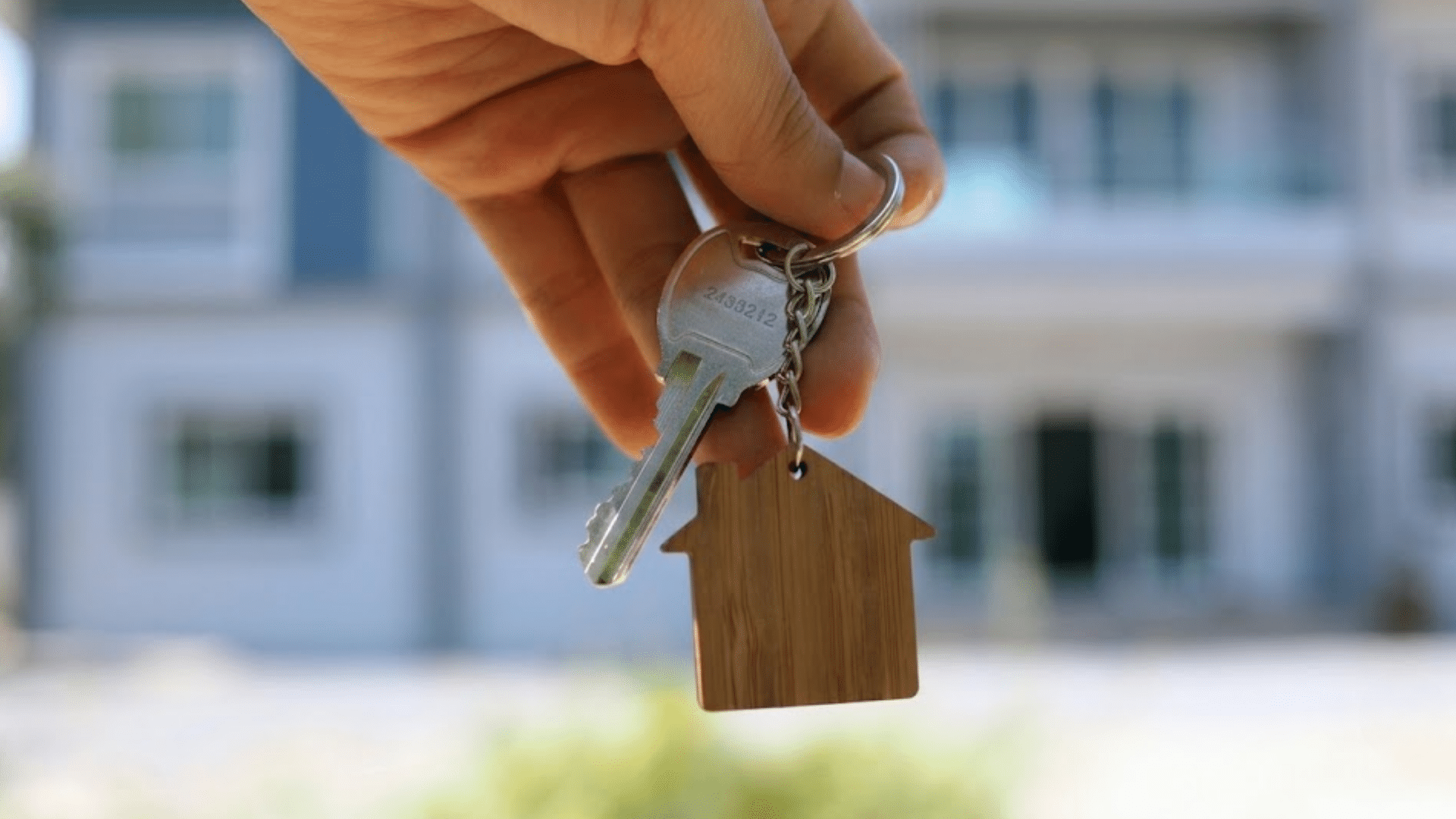 hand holding a house shaped keychain with a metal key in front of a residential building symbolizing home ownership