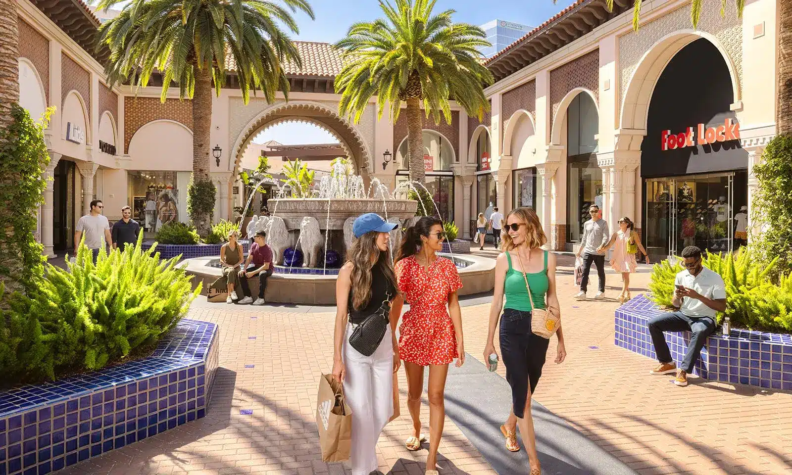 Three women walking through outdoor shopping plaza with palm trees and fountain in sunny weather