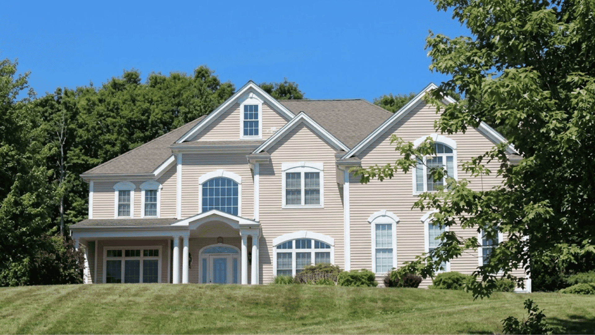 large beige two story house with white columns arched windows a wide front lawn and trees under a clear blue sky