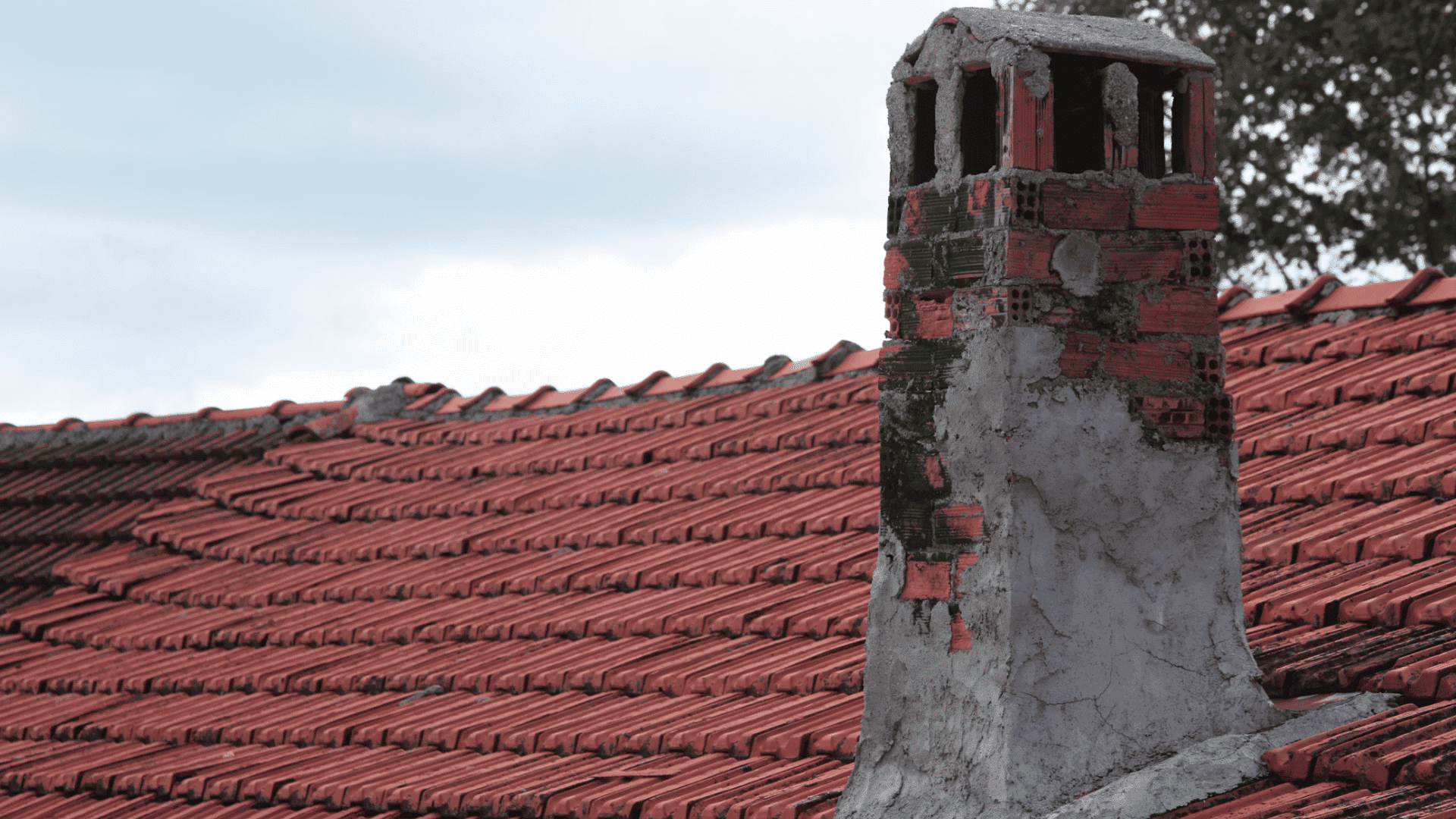 old brick chimney rising from a weathered red tile roof with cloudy sky and trees in the background