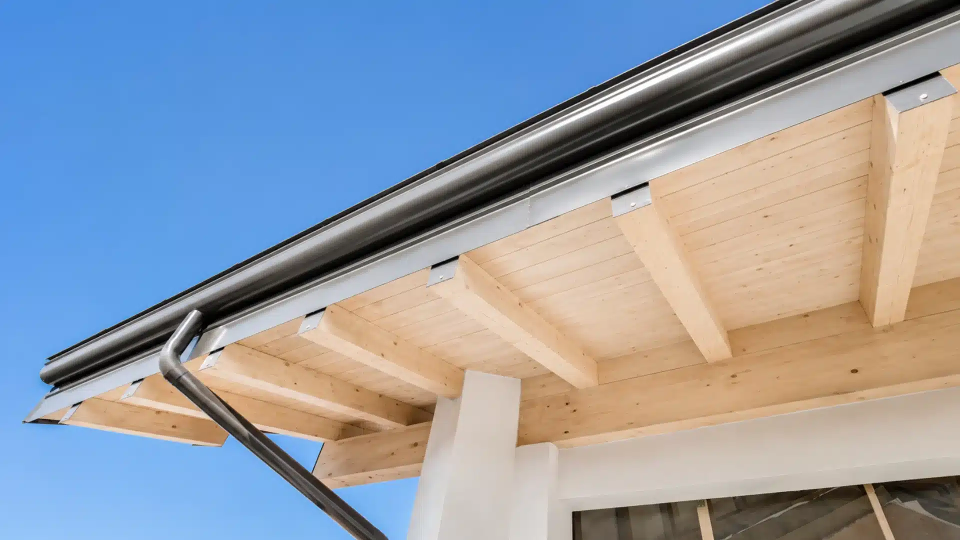Modern house eave with wooden soffit, metal gutter, and downspout against a blue sky.