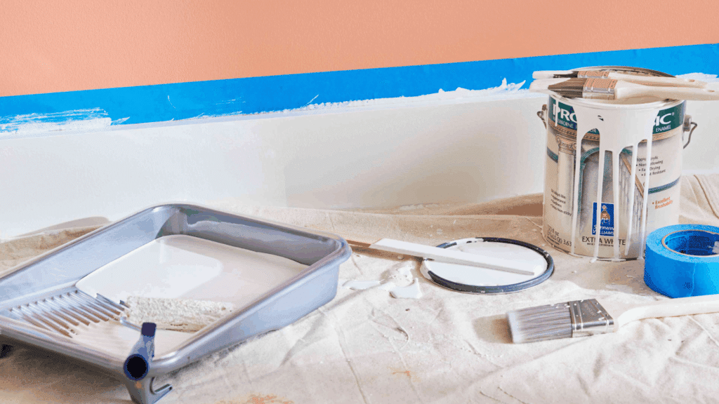 What Is Enamel Paint: Water-Based vs Oil-Based paint tray with white paint beside brushes paint can and blue masking tape in front of a wall being freshly painted