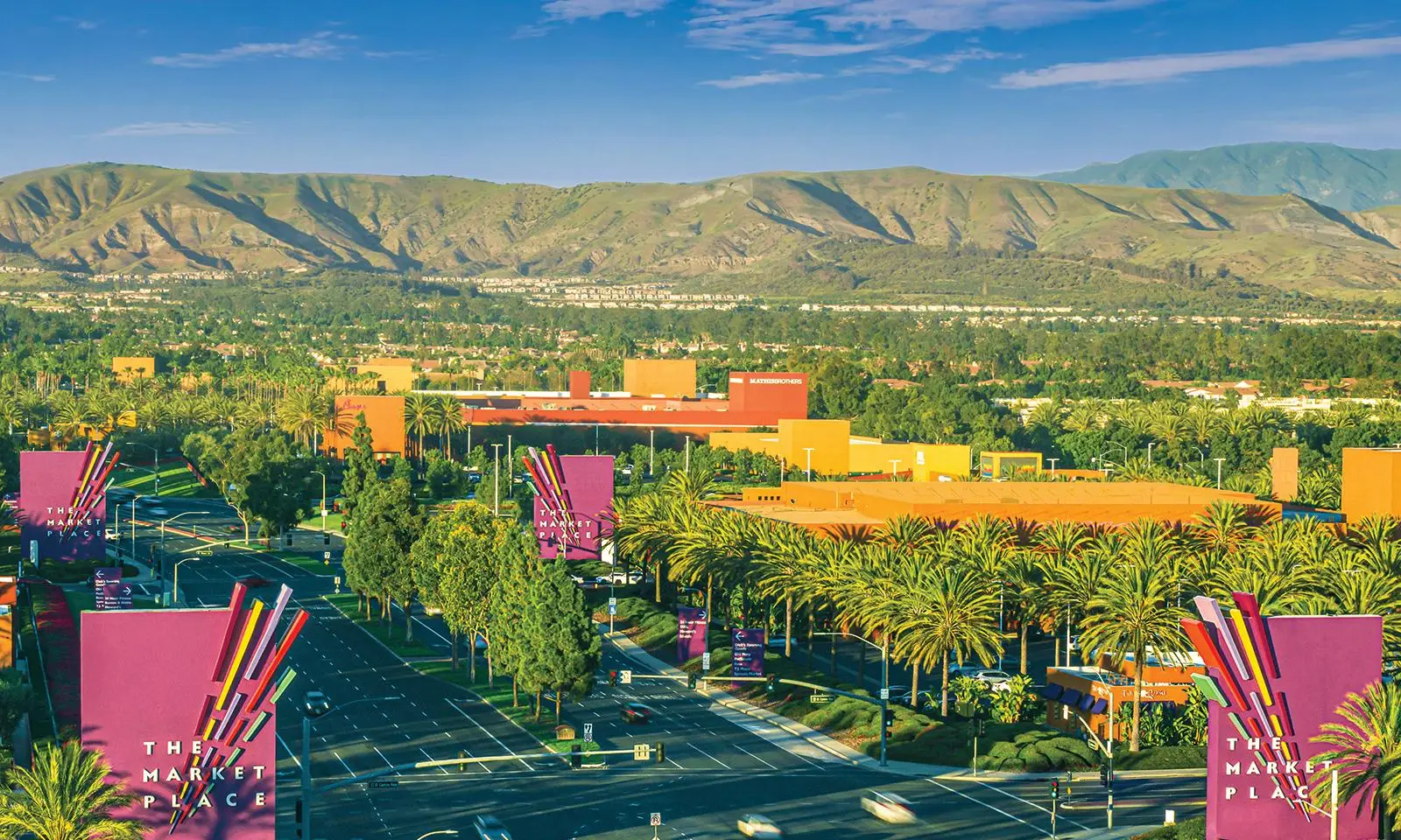 Aerial view of shopping center with colorful signs and lush greenery under clear blue sky