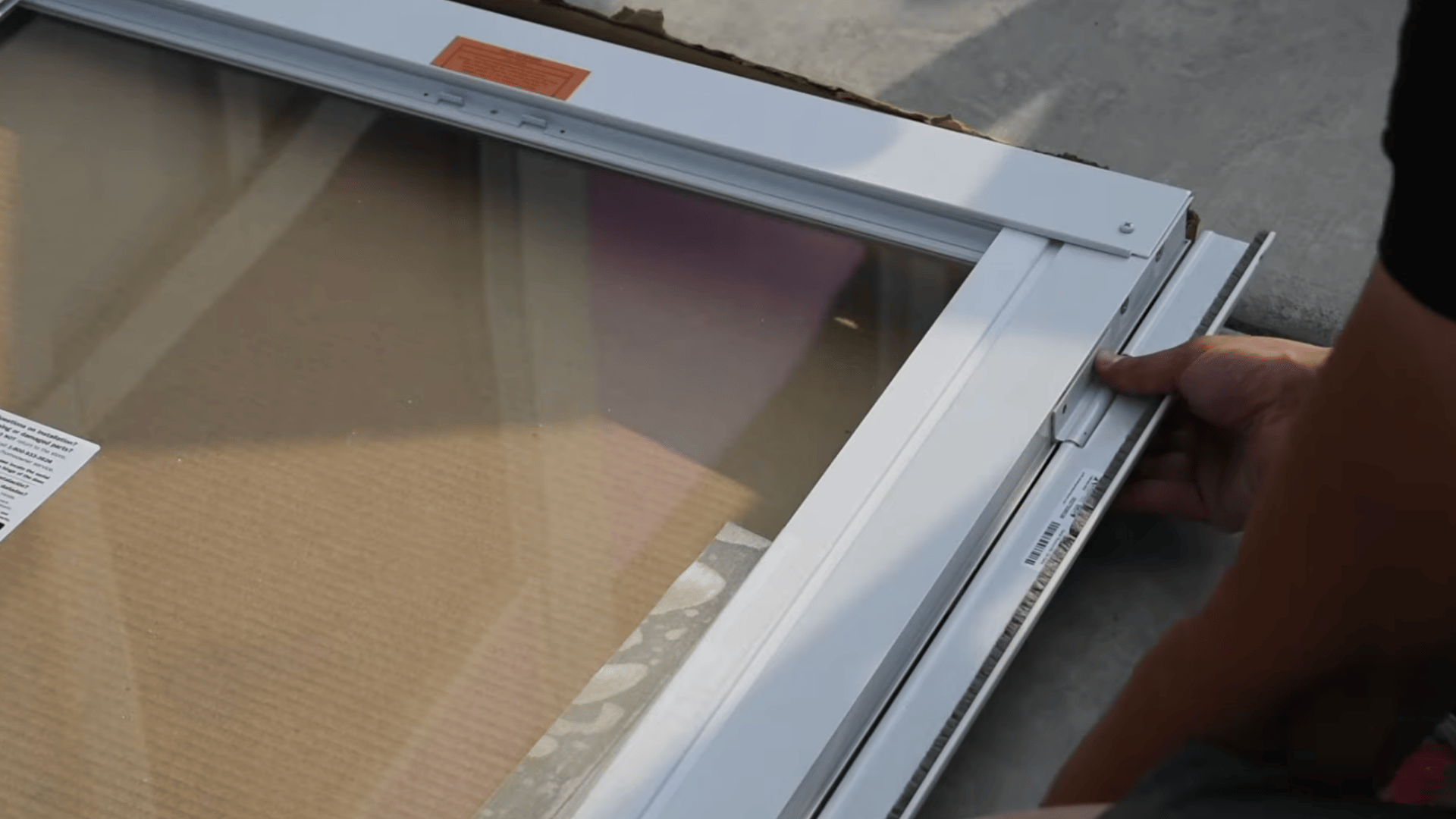 person aligning a white storm door frame on a glass door panel while preparing the parts for installation on a flat surface