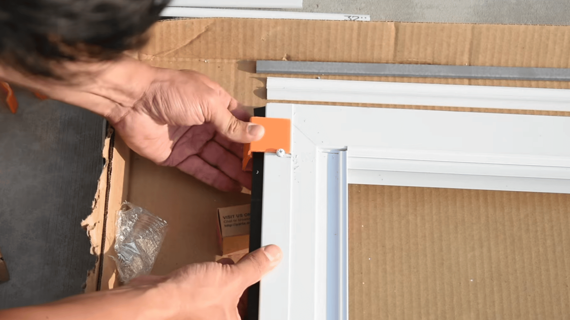 person attaching an orange corner clip to a white storm door frame while assembling the door components on cardboard surface