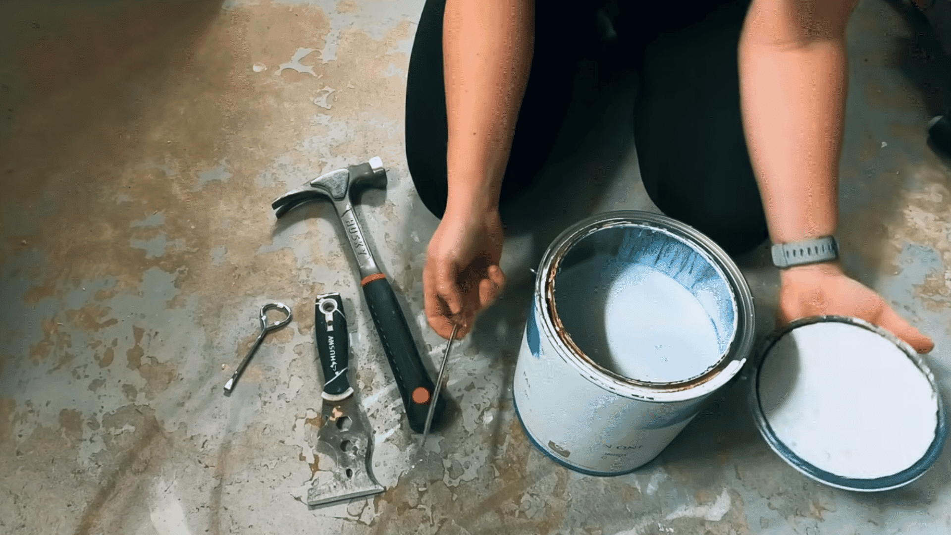 person holding the removed paint can lid beside an opened can of white paint on the floor