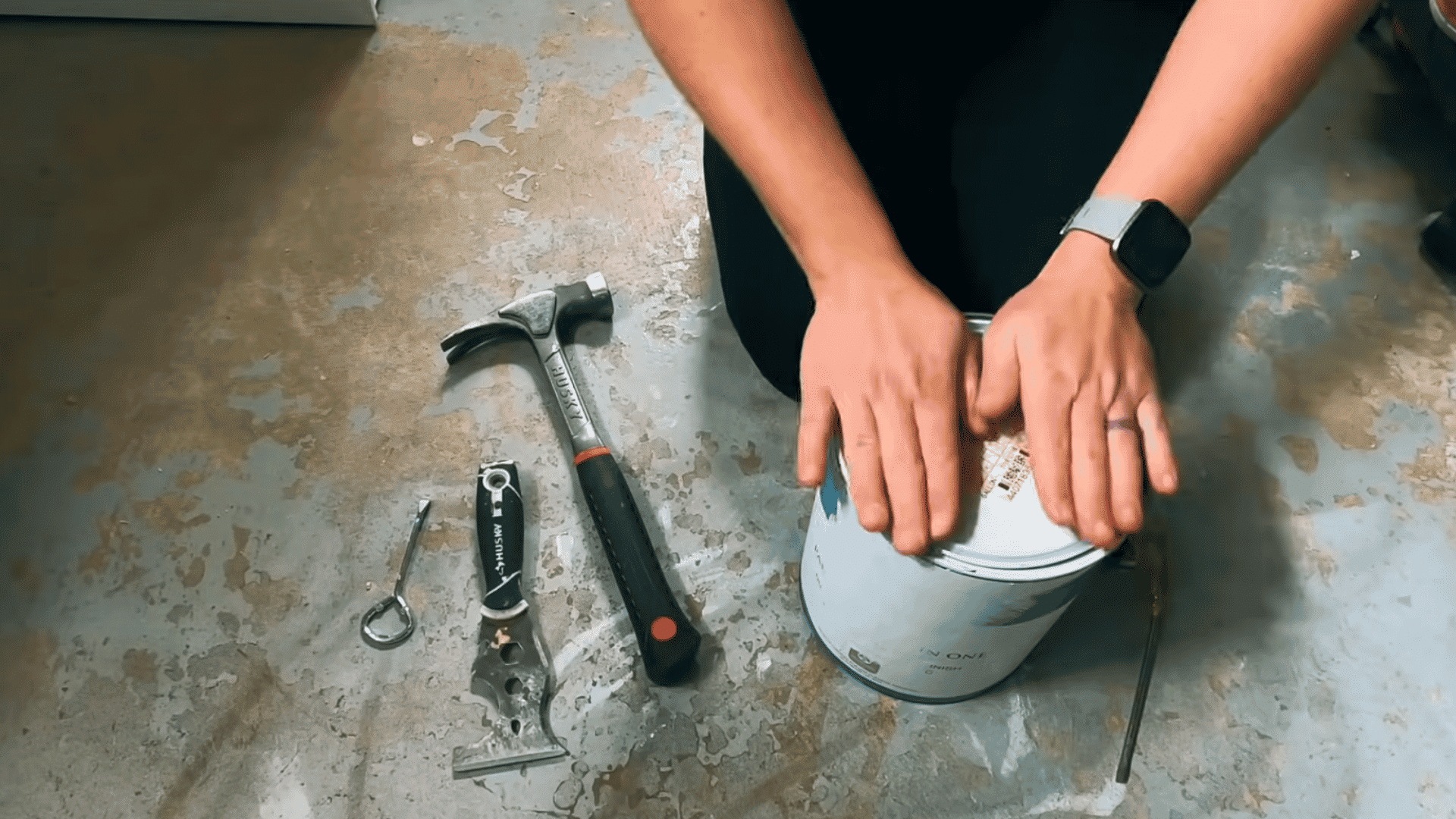 person pressing down on a paint can lid beside a hammer scraper and paint can opener on a concrete floor