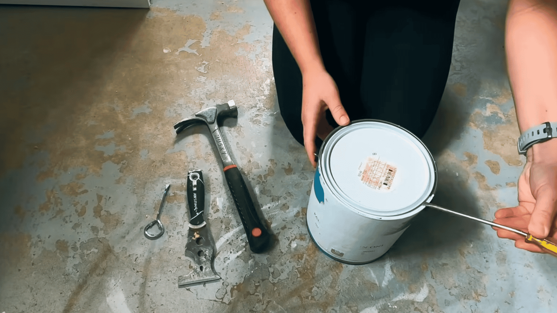 person prying open a paint can lid with a flathead screwdriver beside painting tools on the floor