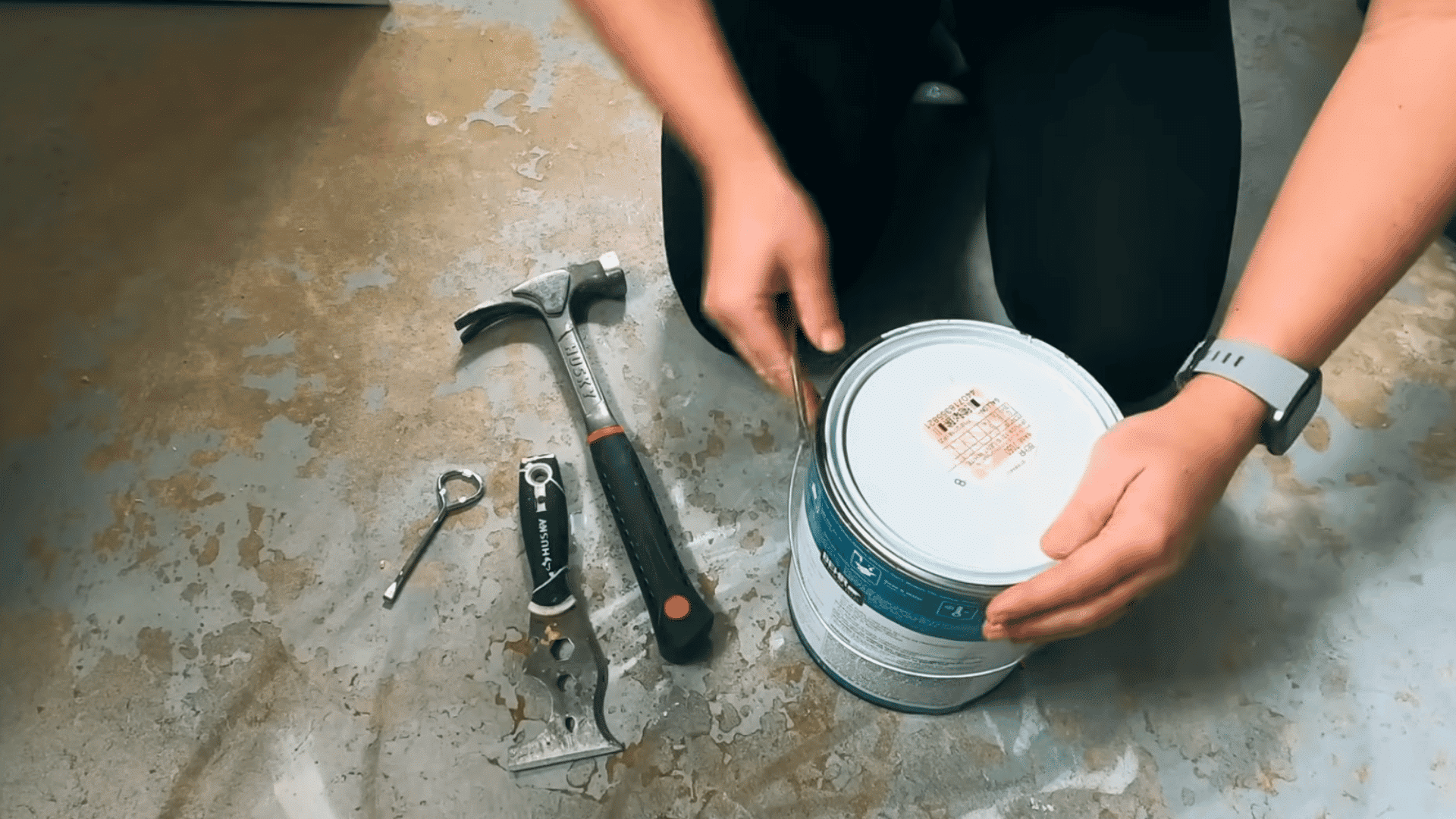 person prying open a paint can lid with a flathead screwdriver beside painting tools on the floor