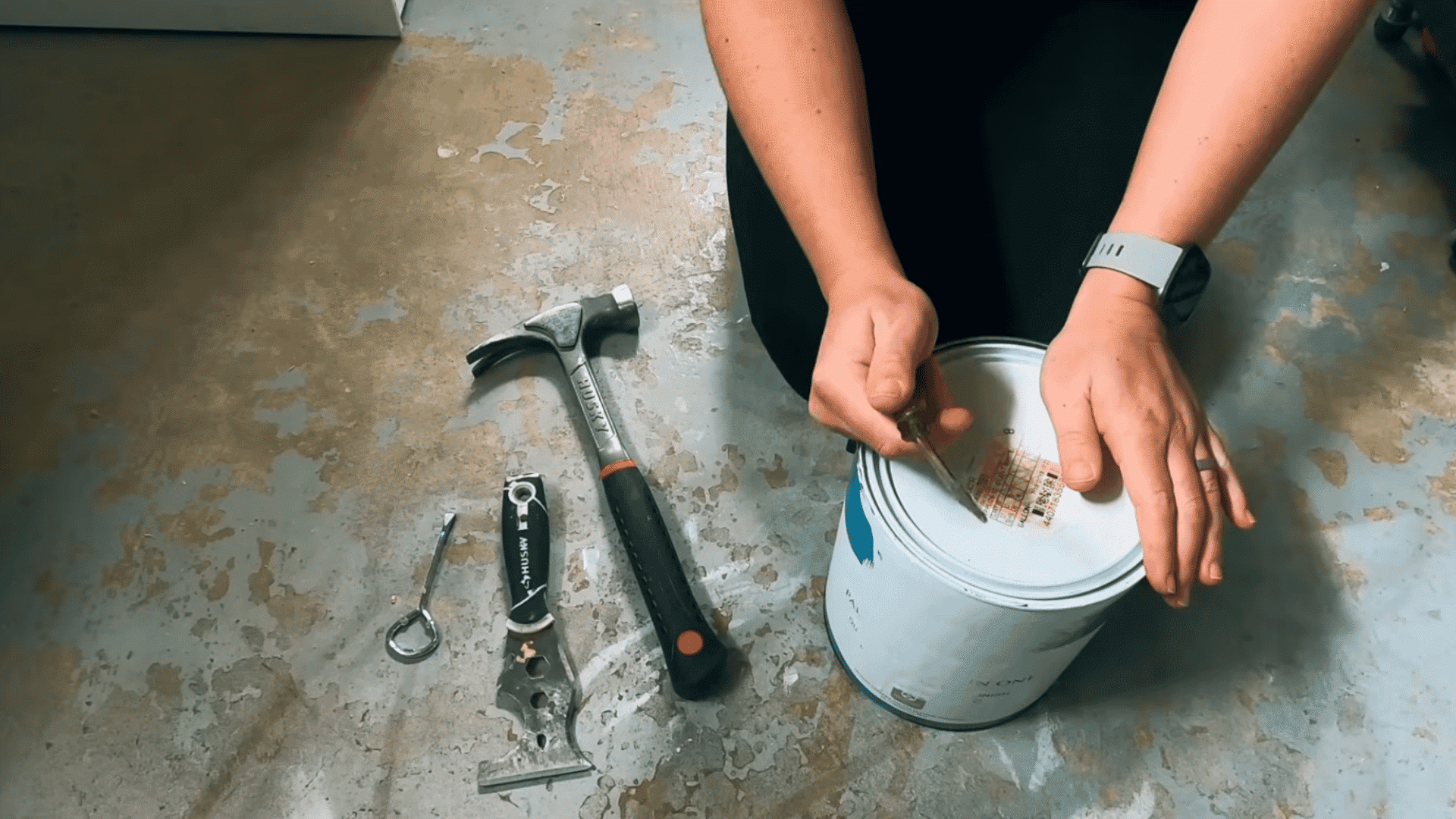 person using a flathead screwdriver to lift the lid of a paint can with tools nearby on the floor