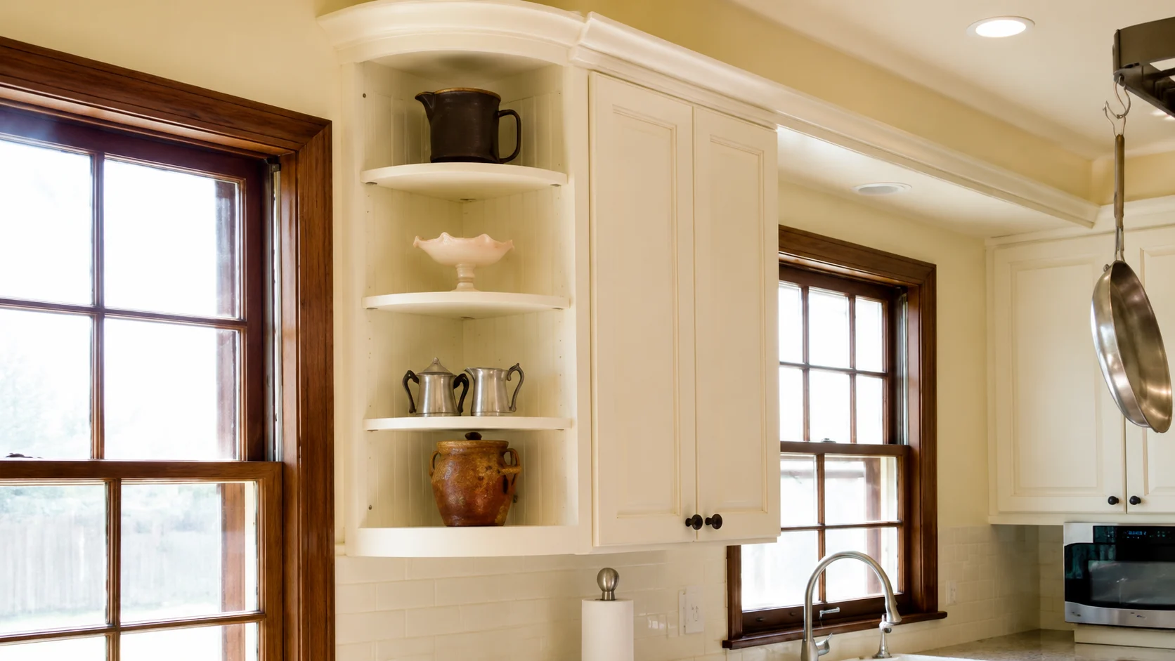 White kitchen with corner open shelves displaying ceramic decor, cream cabinets, wood-trimmed windows, and warm natural lighting.