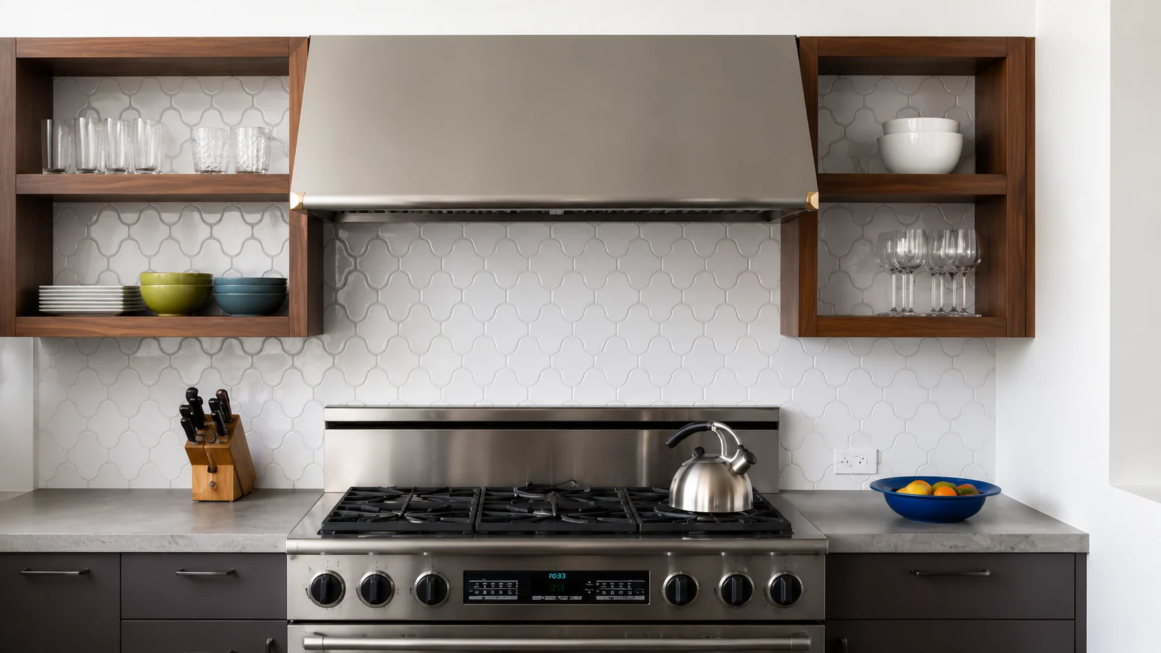 Modern kitchen with wooden open shelves, stainless steel range hood, gas stove, white patterned backsplash, glassware, and dark cabinets.