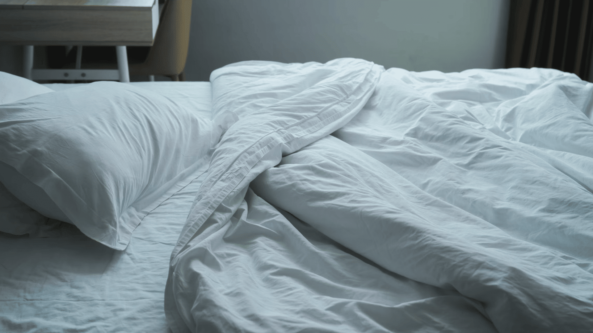 white pillows and a white comforter rest on an unmade bed beside a wooden desk in a softly lit bedroom