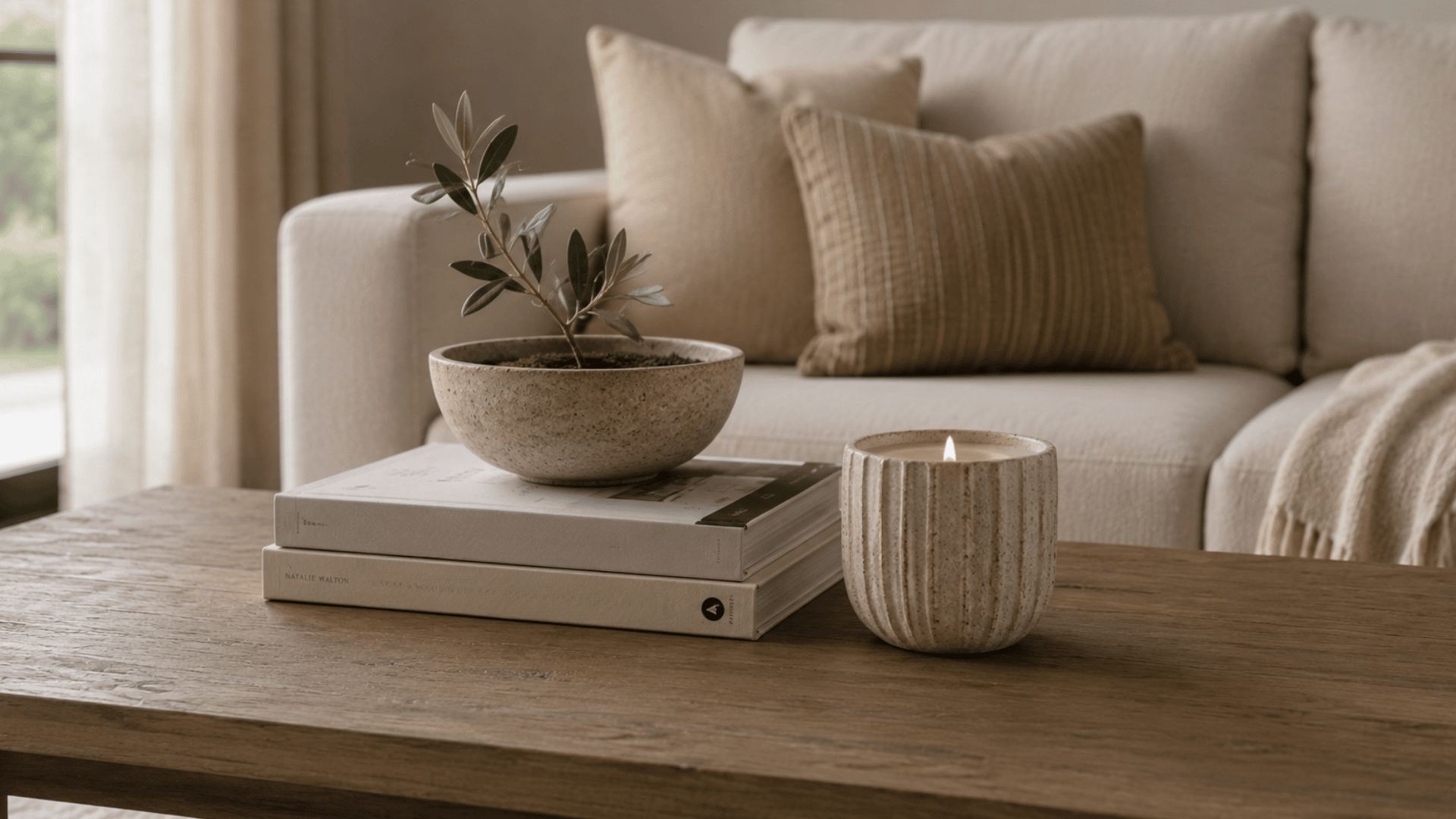 wooden coffee table with potted plant on stacked books and a lit candle in front of a beige sofa with soft cushions and throw