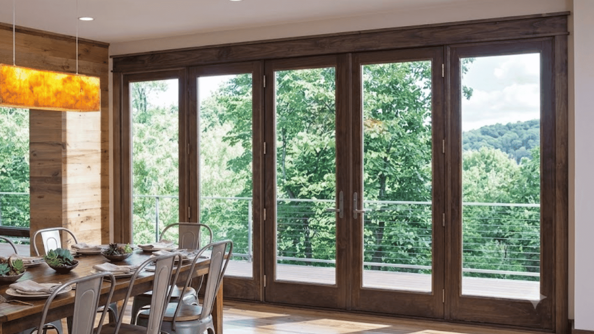 wooden dining room with large glass patio doors overlooking lush green trees beside a rustic dining table