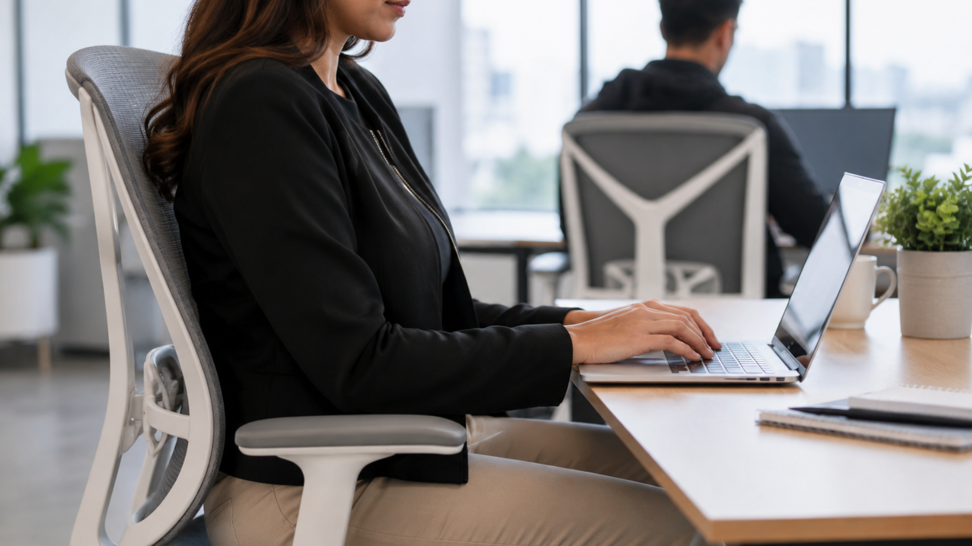 woman in a black jacket and beige pants typing on a laptop while seated in an ergonomic office chair