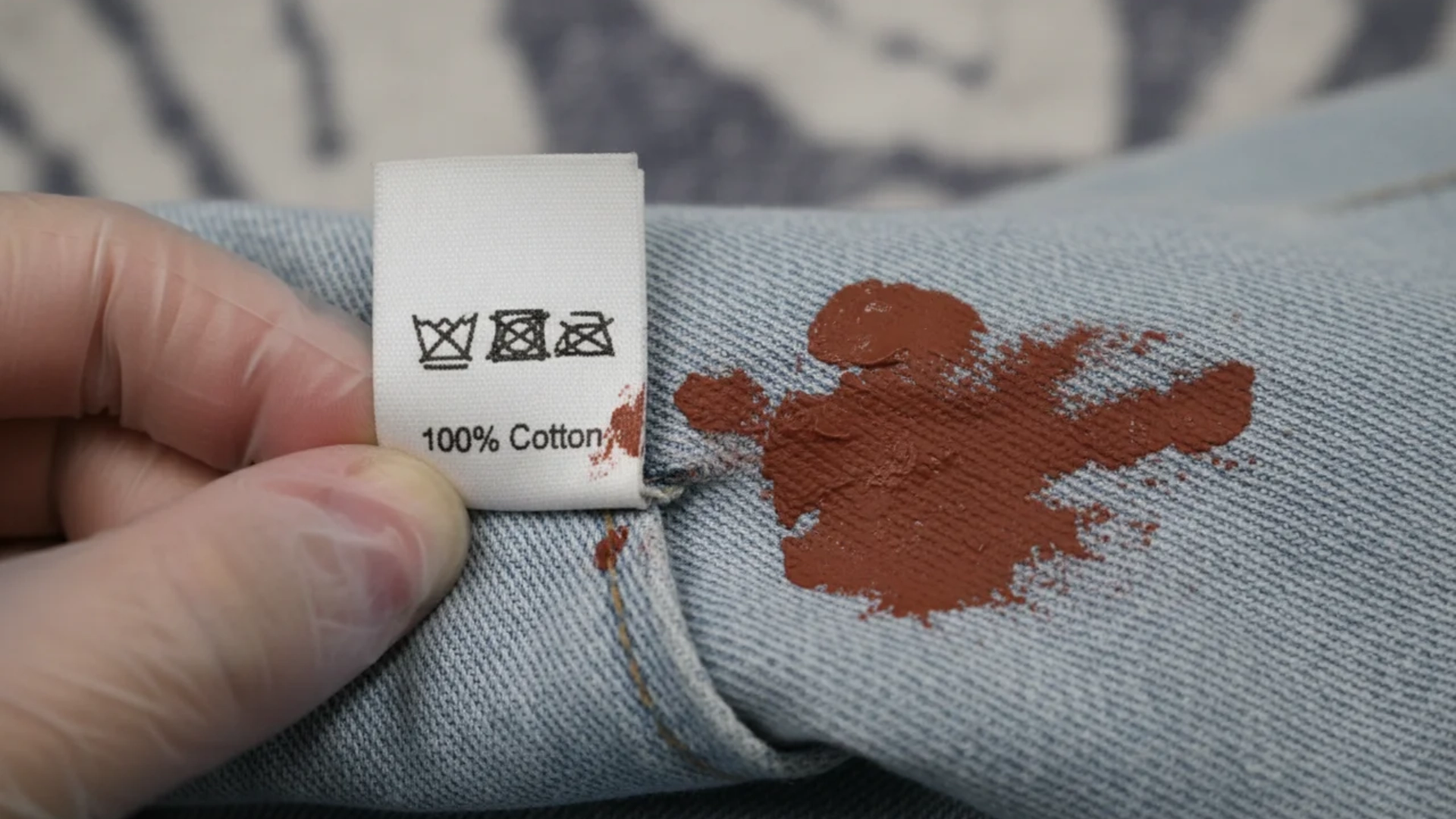 A hand holding a cotton clothing label near a brown paint stain on denim.