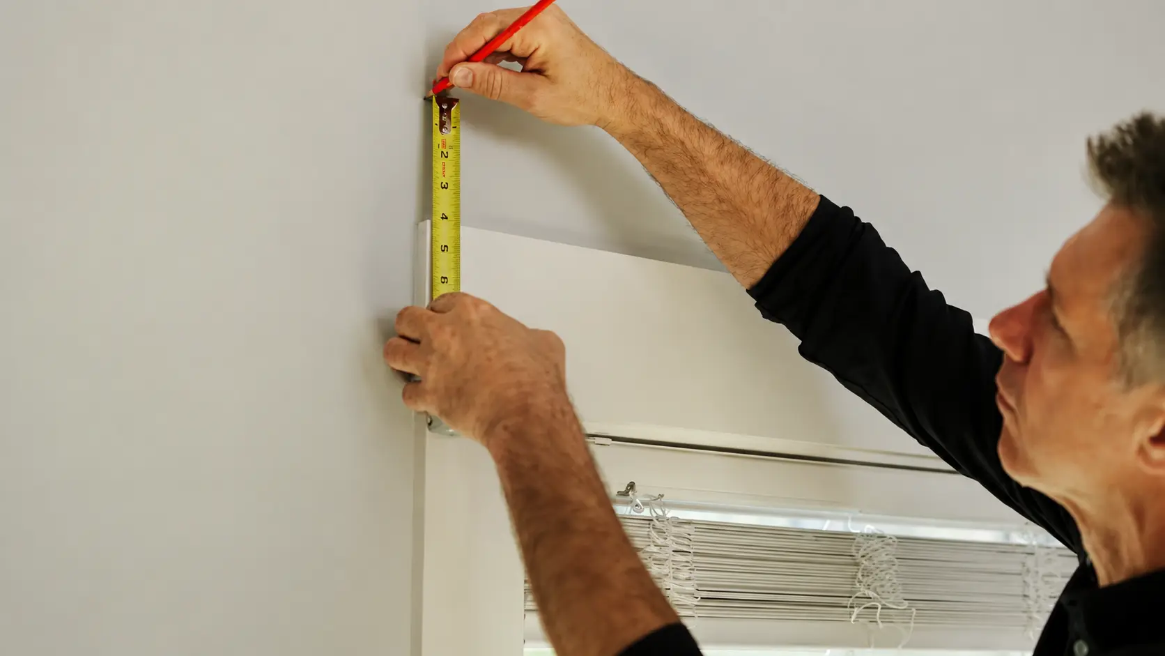 Person measuring above a window with a tape measure and pencil to mark curtain placement