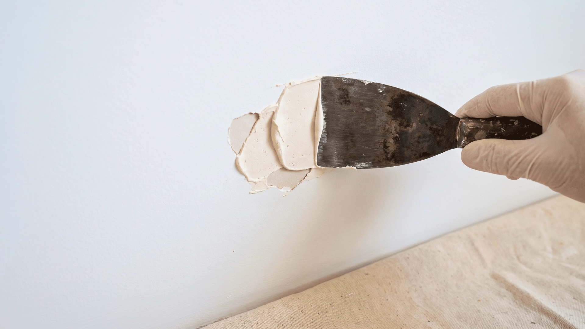 Putty knife applying filler to repair a damaged section of a white wall before painting.