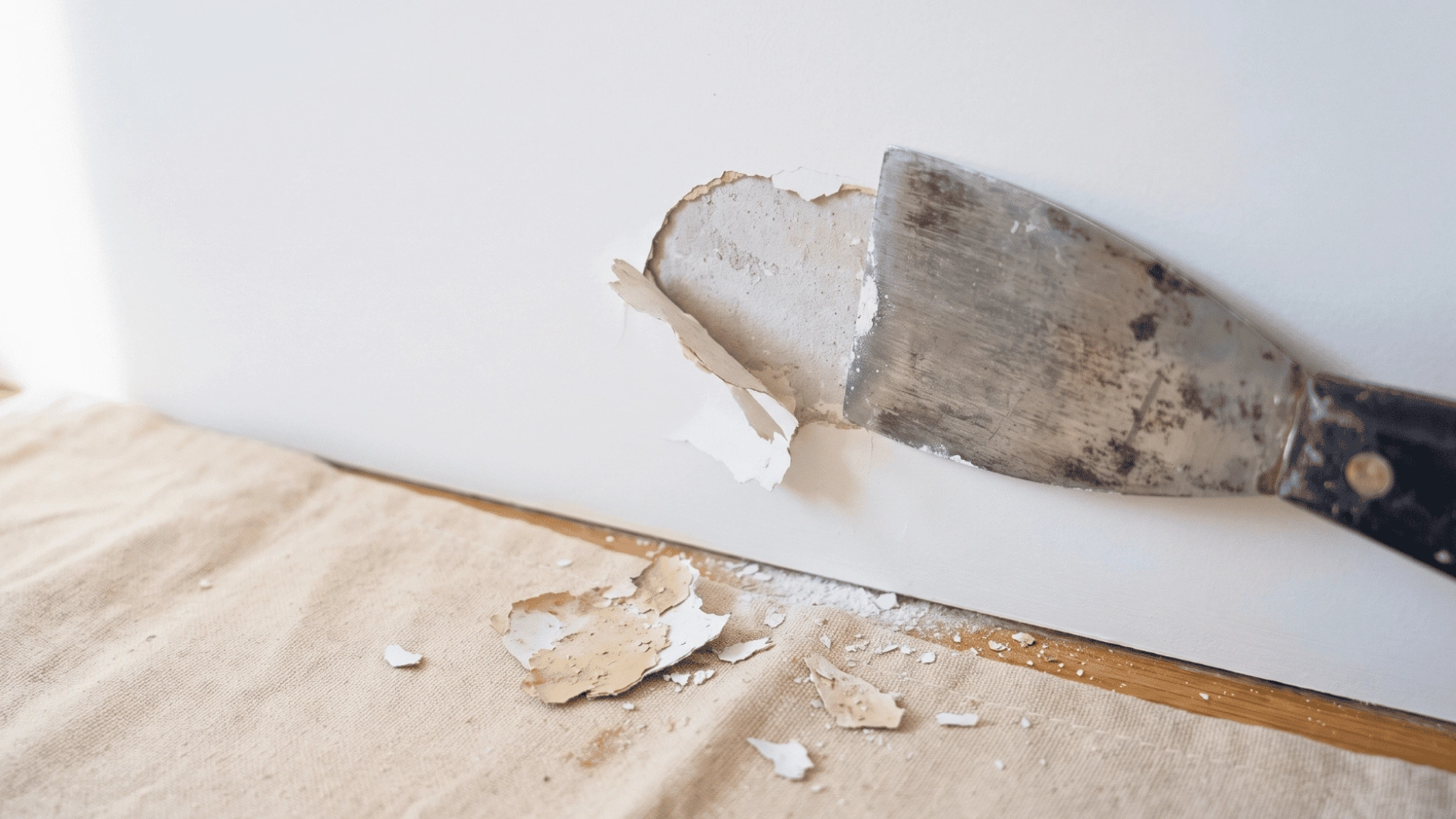 Putty knife removing peeling paint from a white drywall wall with paint chips on a drop cloth.