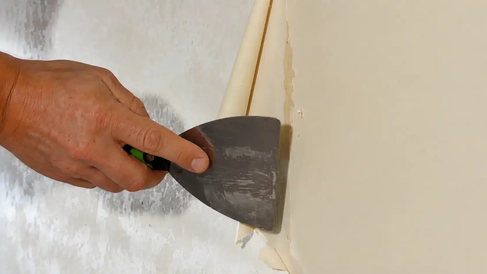 Hand using a putty knife to scrape softened wallpaper glue residue from a wall surface during removal