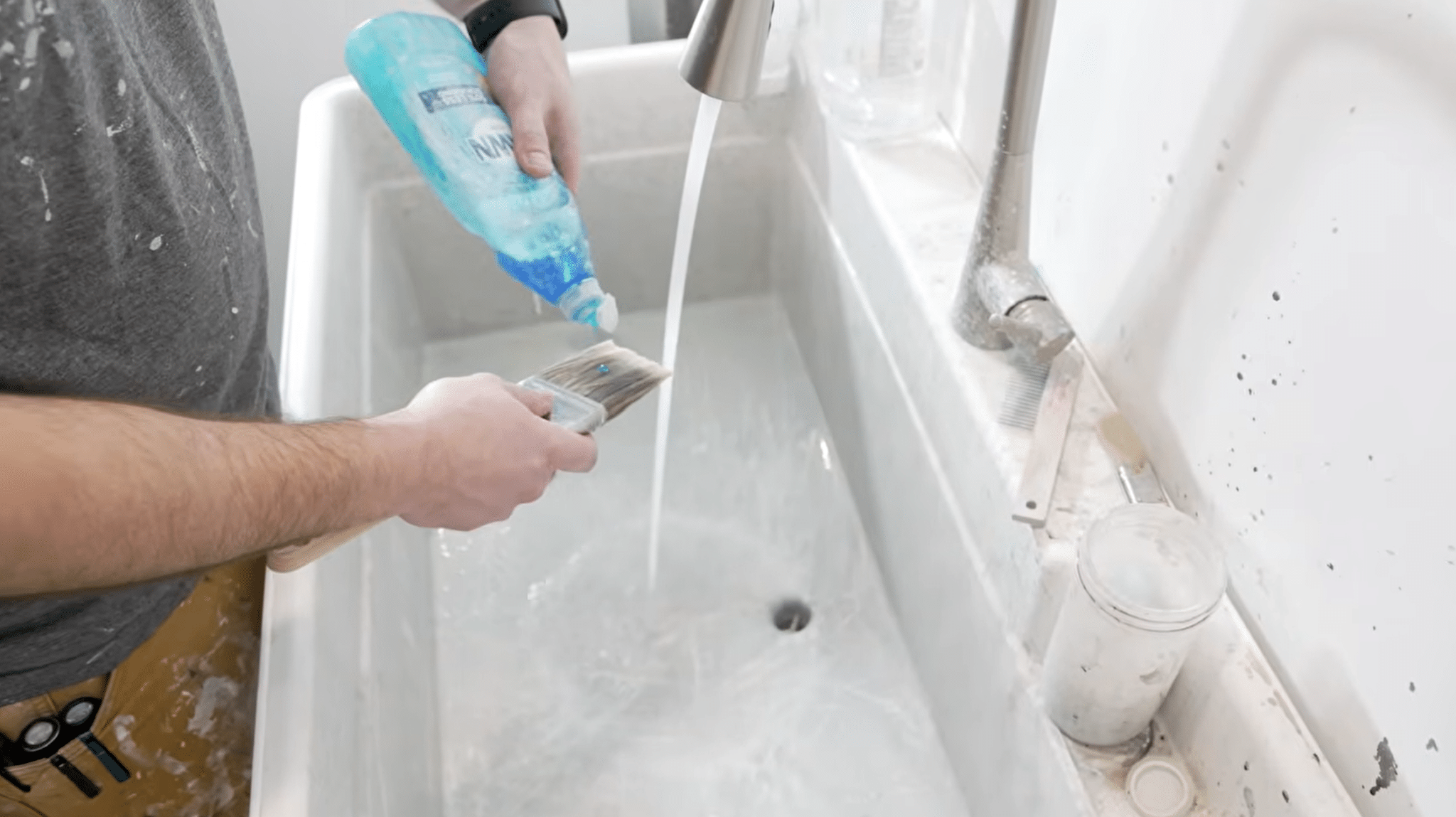 Applying dish soap to a paint brush while rinsing it under running water in a sink.