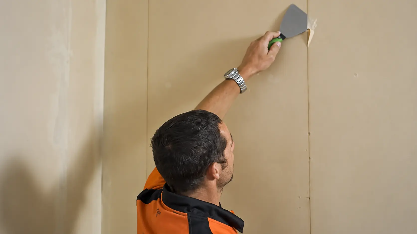 Person using a putty knife to scrape wallpaper glue residue from a beige wall during removal work