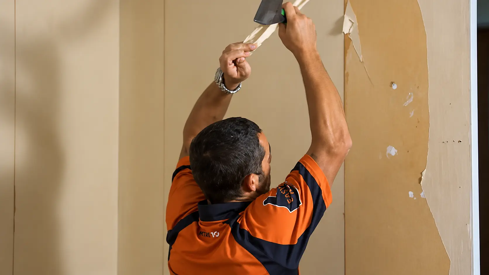 Person scraping old wallpaper from a beige wall using a putty knife during a home renovation project