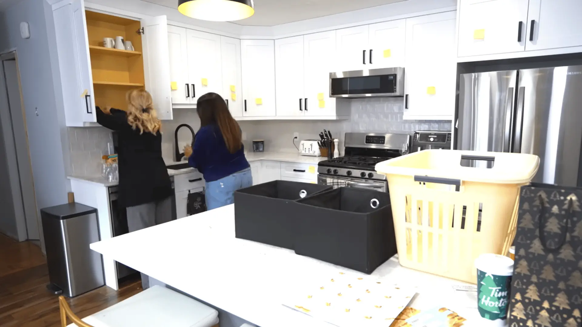 Two women empty kitchen cabinets and relocate items into their planned storage zones using baskets and bins.