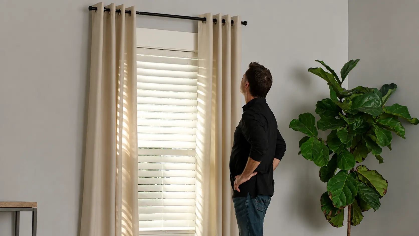 Man checking beige curtains hanging from a black curtain rod beside a bright window