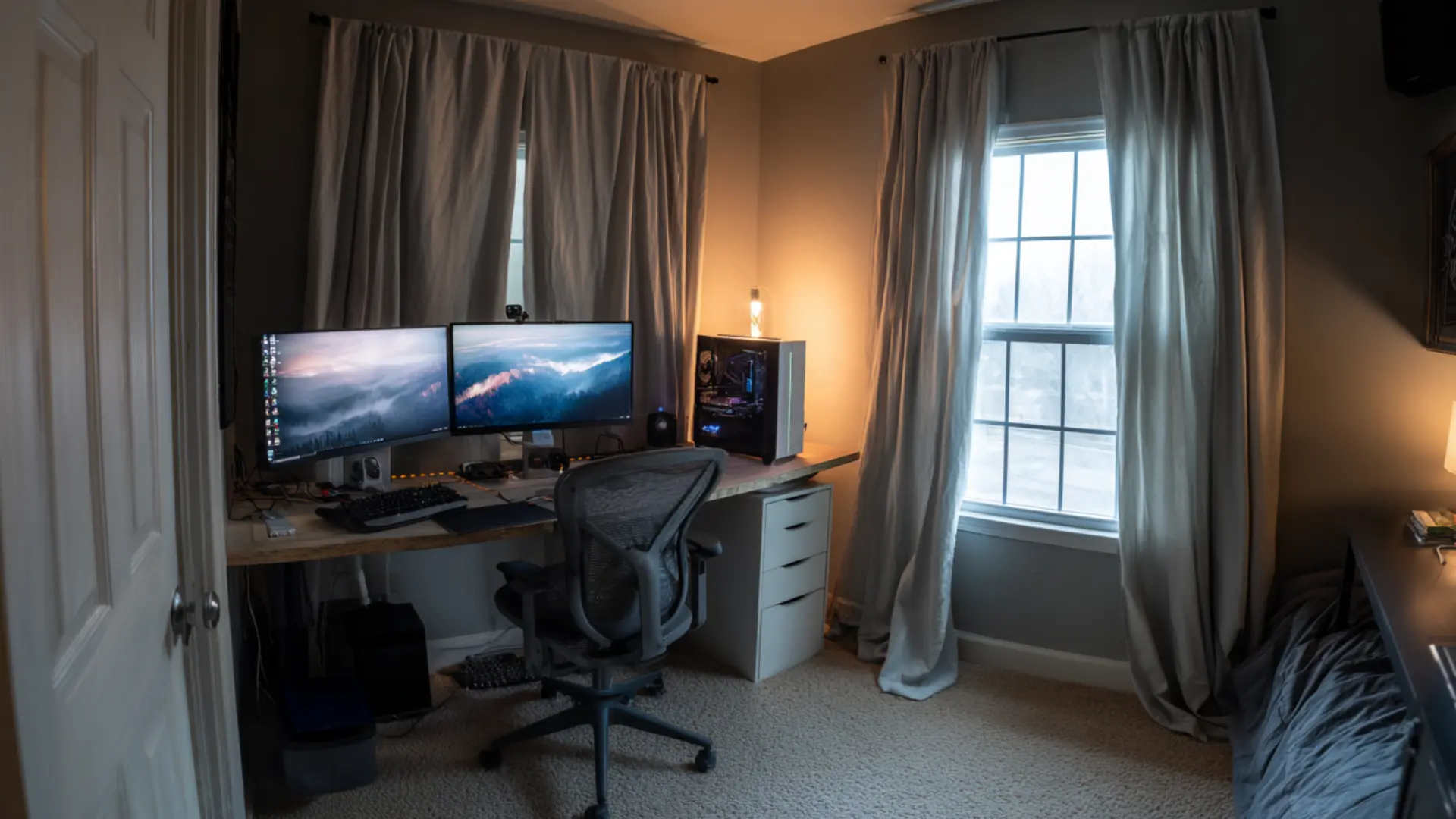 den gaming setup with a long desk two wide monitors a mesh task chair a tower pc and a curtained window