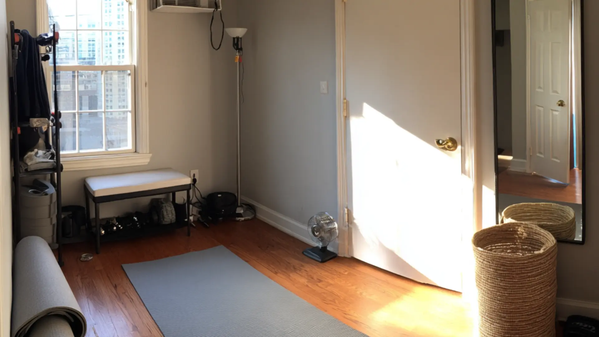 den home gym with a grey yoga mat on wood flooring a padded bench weight rack floor lamp and tall mirror