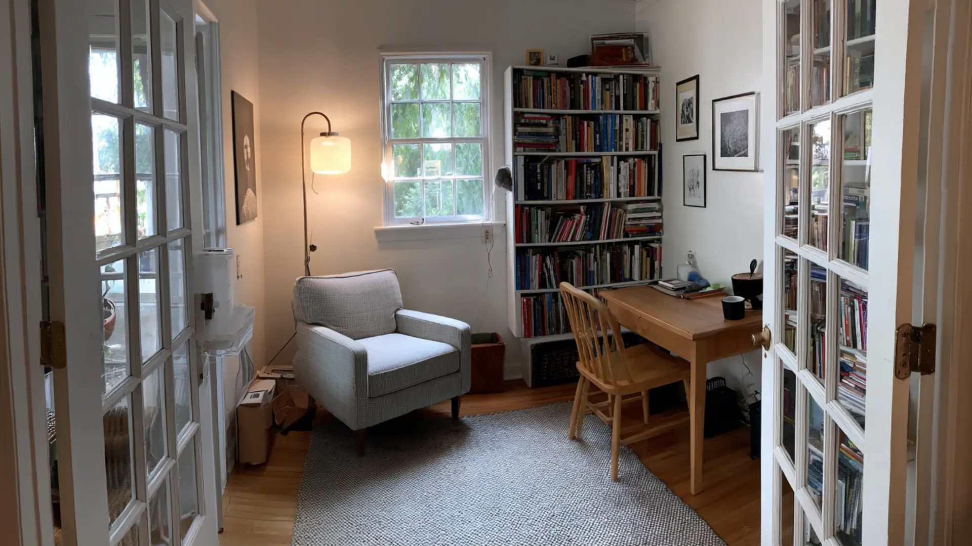 compact den with a grey armchair arc floor lamp wooden writing desk crowded bookshelves and a single window