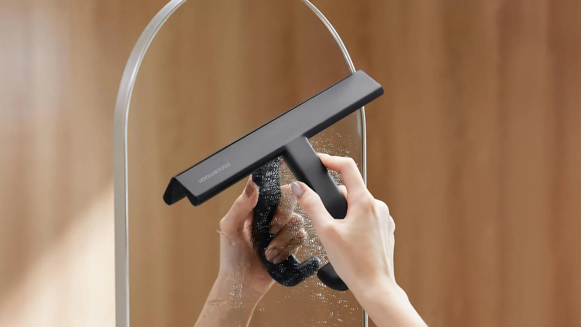 Hands cleaning a mirror with a black squeegee in a softly lit interior setting